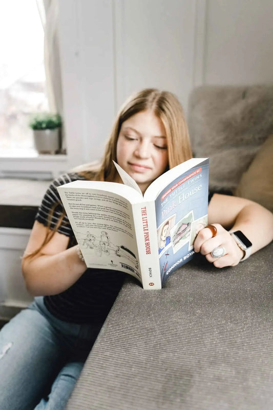 Child reading a small book on a couch, illustrated cover with a pink house