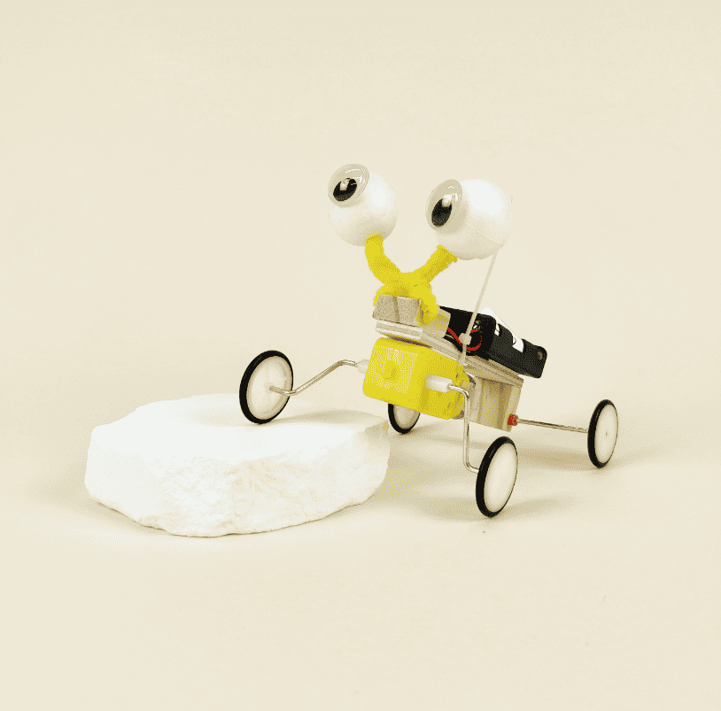 Bug robot DIY kit on a small white pad: googly eyes, yellow antenna, and wheeled body