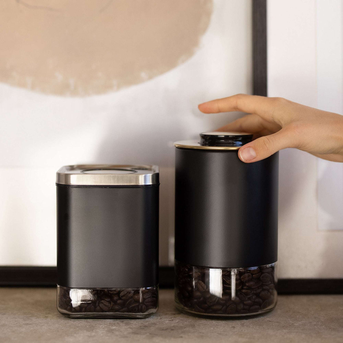 Pair of round matte black storage containers with stainless lids and clear glass bases