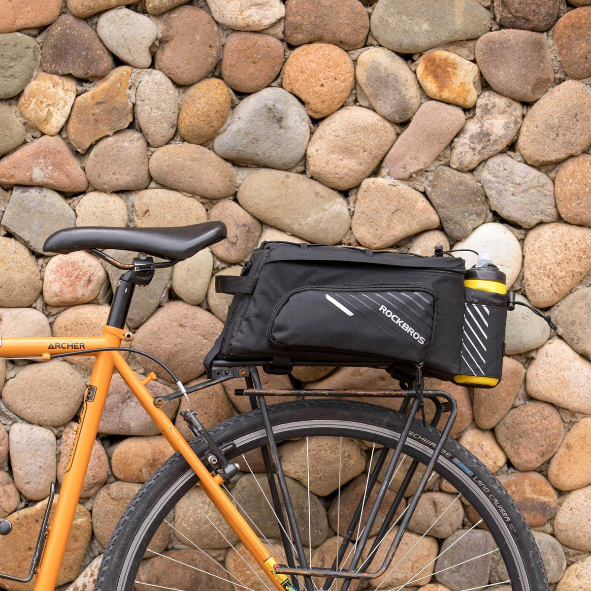 Black bike rack bag with side bottle holder and zip pockets on bicycle near stones