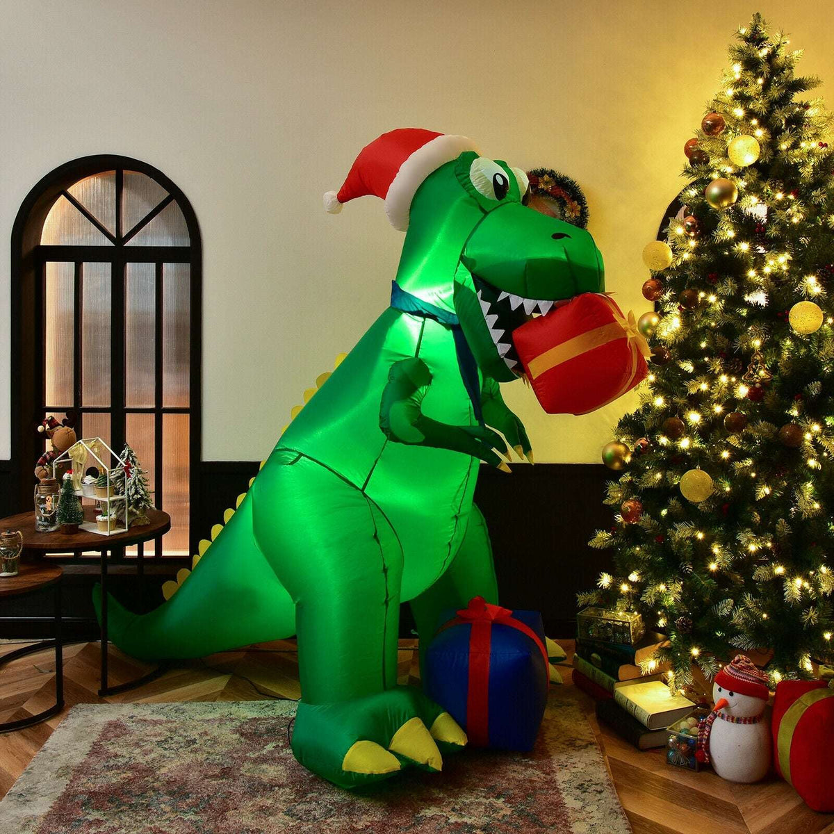 Green Christmas inflatable dinosaur holding a gift beside a lit Christmas tree