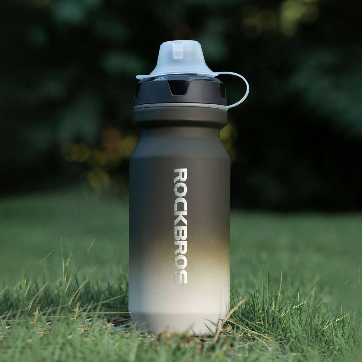 Black-to-gray gradient bike water bottle with flip-top lid standing in grass